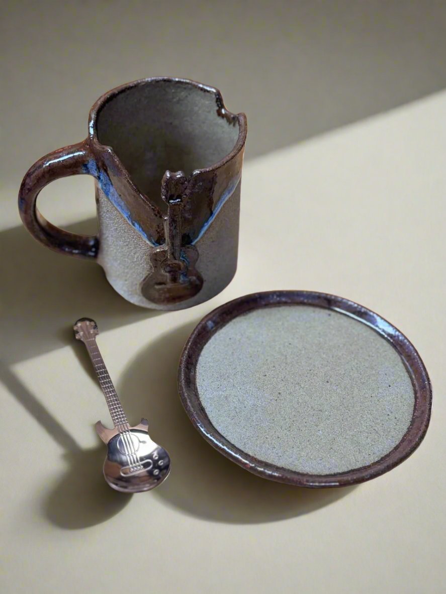 La abertura de la taza en forma de cuello y el adorno de guitarra se muestran de manera prominente. El platillo a juego y la cuchara en forma de guitarra completan el conjunto. La disposición está colocada sobre una superficie de madera, enfatizando la calidad artesanal de las piezas.